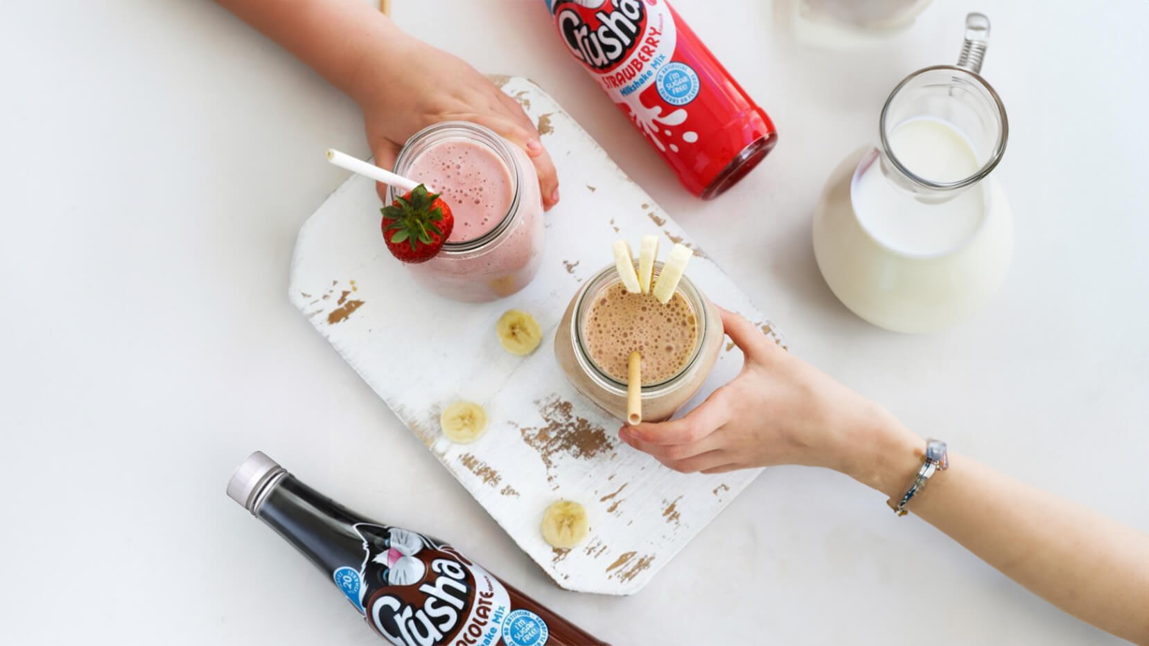 Strawberry and banana milkshakes in glass jars with fresh fruit on a countertop alongside Crusha Milkshake bottles, with two chidlren's hands reaching to grab them.