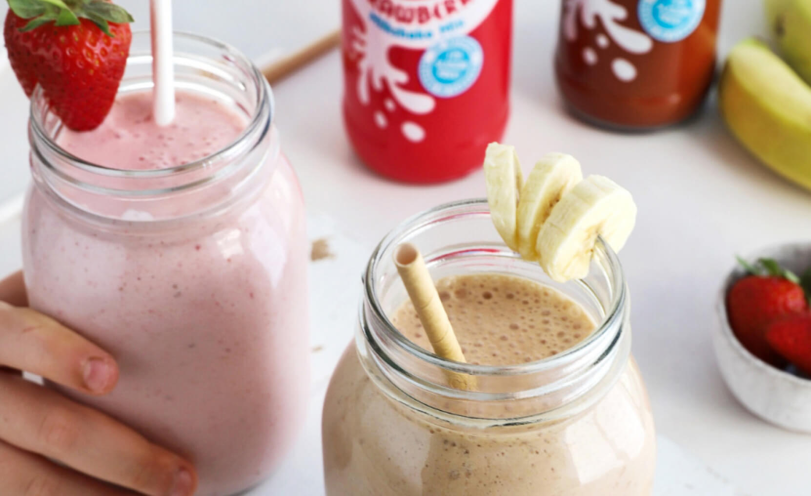 Banana and Strawberry milkshakes in glass jars with added fresh fruit, and Crusha milkshake bottles in the background.