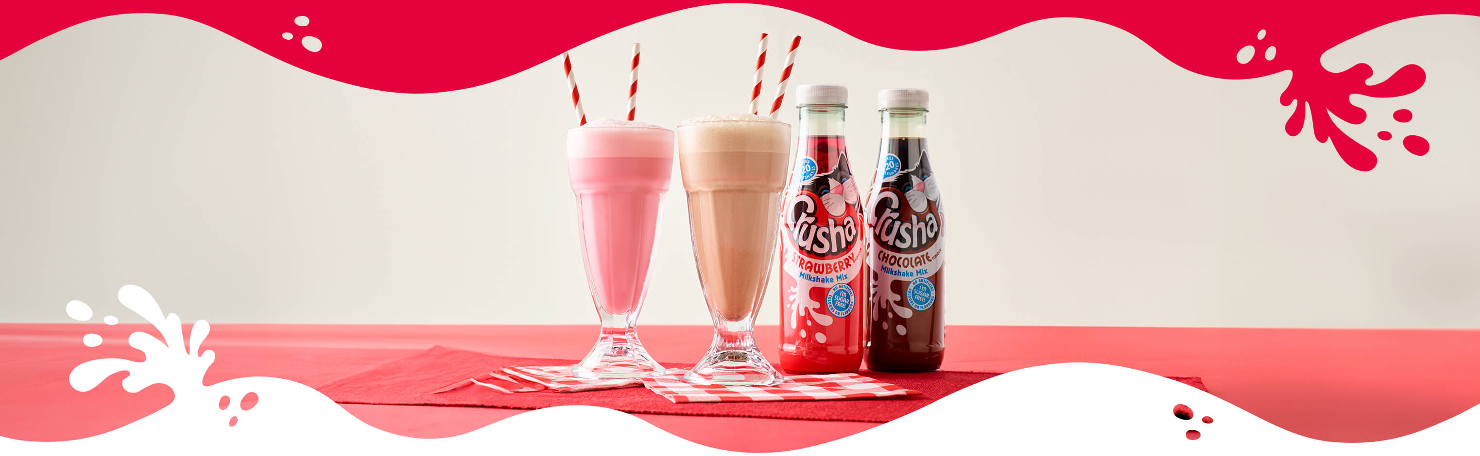 Crusha Milkshake Mix bottles alongside two milkshakes in tall glasses with stripy straws.